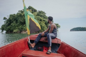 Man sitting in a red wooden boat with a Jamaican flag, looking at a green island 